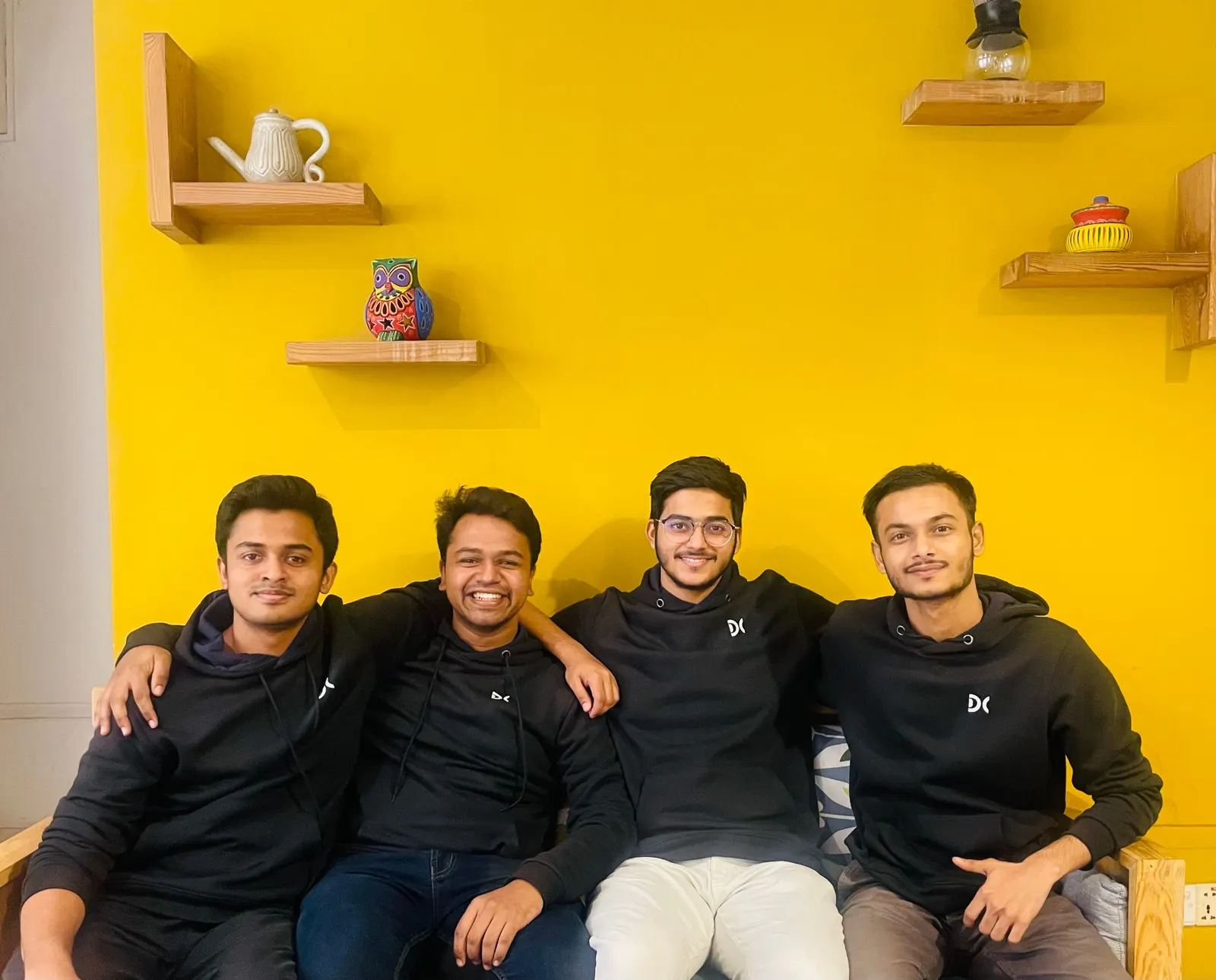 Four Devs Core engineers in company hoodies on a sofa in front of a yellow wall